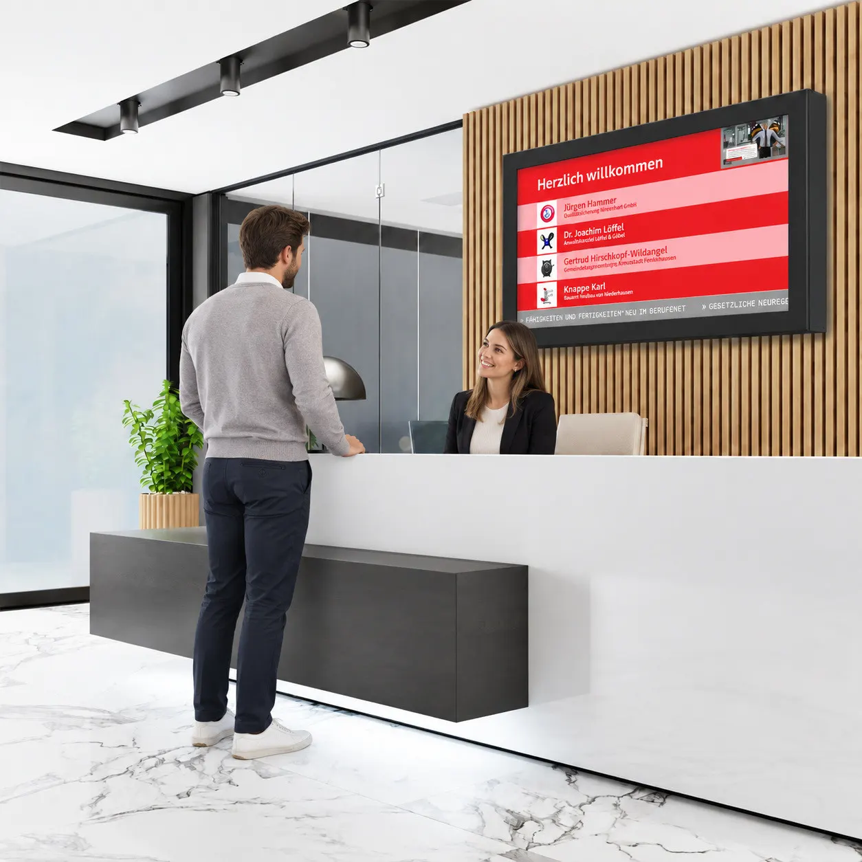 Besucher am Empfangstresen eines Unternehmens; über dem Tresen zeigt ein großes Wand-Display im Querformat die Besucherbegrüßung und die Termine des Tages.